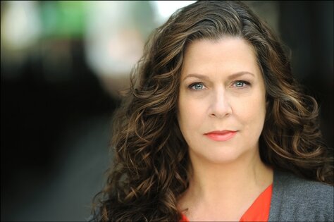 Erin's headshot in which she wears a bright red shirt and a gray cardigan sweater. Her long, gently curled brown hair falls past her shoulders.